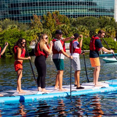 Six people paddleboarding together on a large board in a waterway.