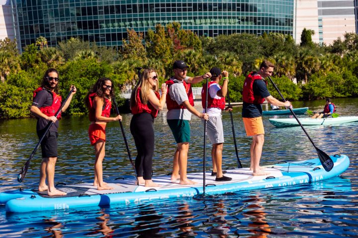 Six people paddleboarding together on a large board in a waterway.