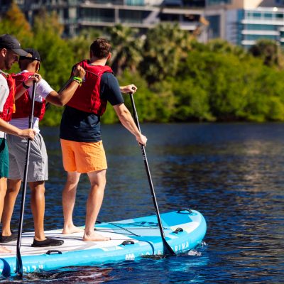 Group paddleboarding on a river with buildings in the background.