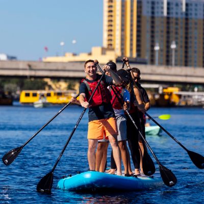 Four people paddle on a large board in an urban waterway.