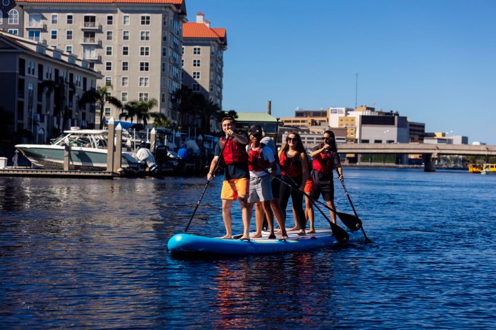 Group paddling on a large paddleboard in a city waterway.