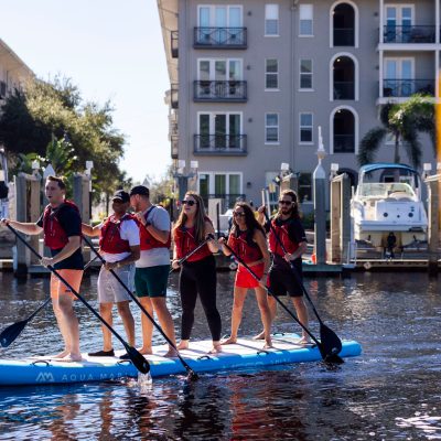 Six people paddleboarding together on a large blue board in a marina.
