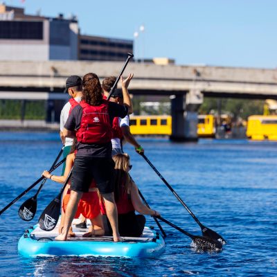Group paddleboarding on a river near a bridge with yellow boats.