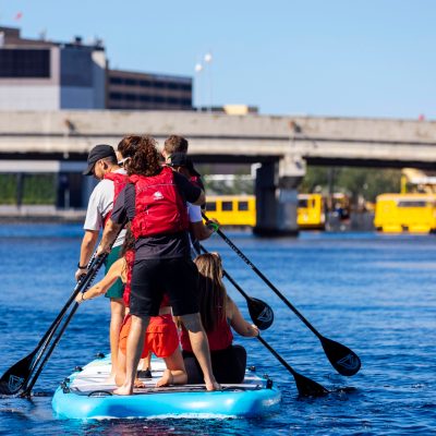 Group of people paddleboarding on a river with a bridge in the background.