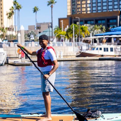 Person paddleboarding on water near docked boats and buildings.