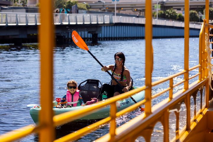 Woman and child kayaking on a river near a yellow railing and bridge.