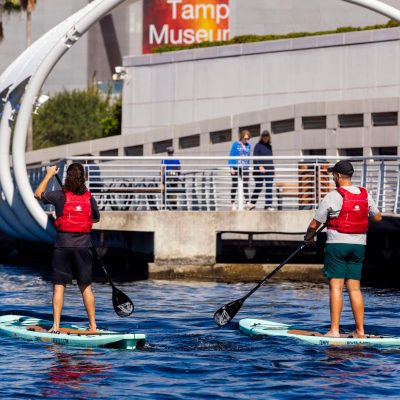Two people paddleboarding near a waterfront building with palm trees.