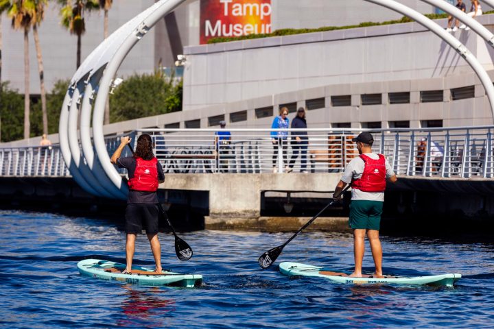 Two people paddleboarding near a waterfront building with palm trees.