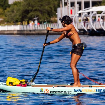 Woman paddleboarding on a river near a city walkway.