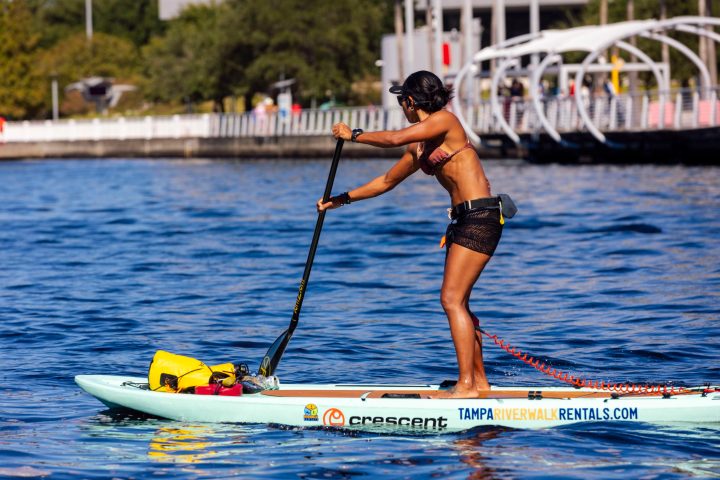 Woman paddleboarding on a river near a city walkway.