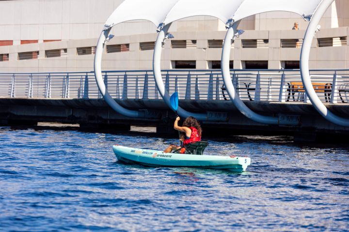 Person kayaking on water near urban bridge with blue paddle.