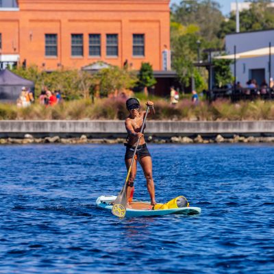 Person paddleboarding on a river, buildings in the background.