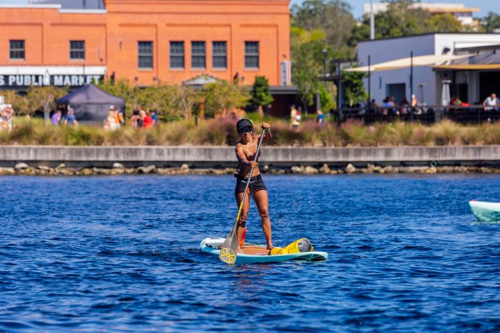 Person paddleboarding on a river, buildings in the background.
