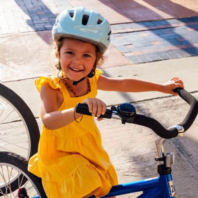 Child in a yellow dress smiles while riding a bike with a helmet.