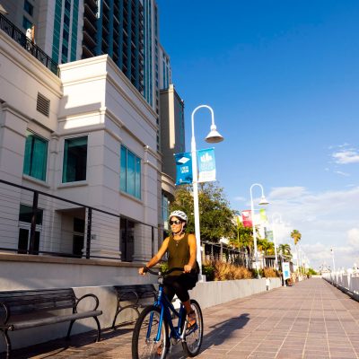 Person cycling on a riverside path near tall buildings on a sunny day.