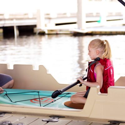 Child in life jacket sits in kayak, smiling at instructor on dock.