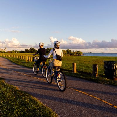 Two cyclists riding on a paved path through a grassy park near the ocean.