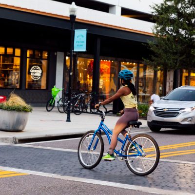 Person on a bicycle crossing a street in front of a cafe and parked cars.