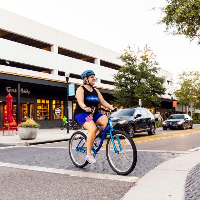Woman cycling on a city street with cars and shops visible.