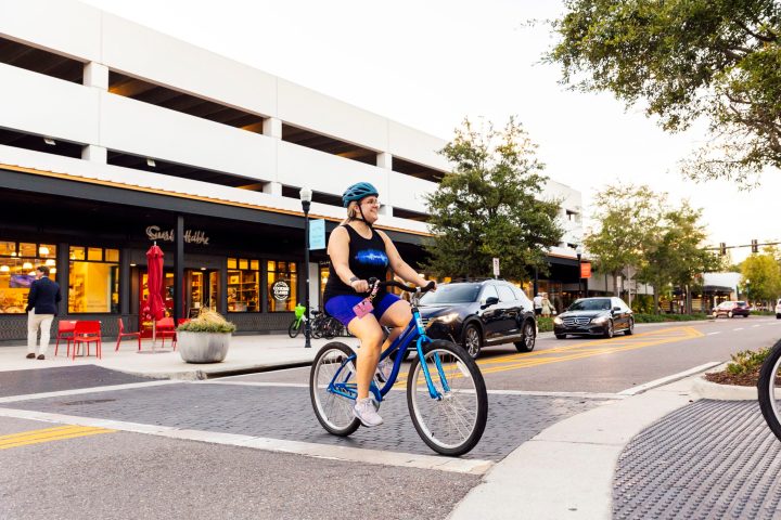 Woman cycling on a city street with cars and shops visible.
