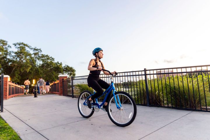 Woman in blue helmet riding a bike on a path with a fence, trees, and people in the background.