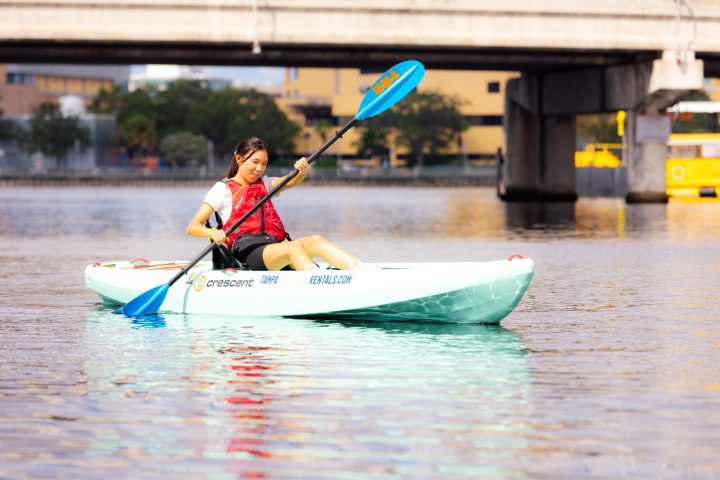 Person kayaking on a calm river under a bridge.