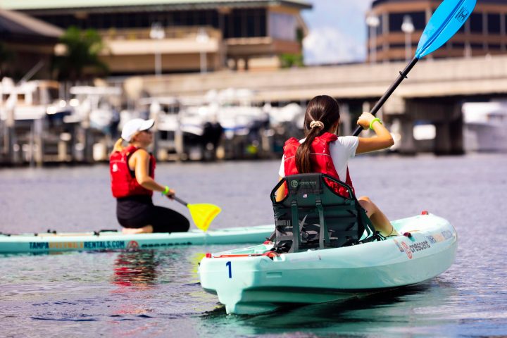 Two people wearing red life jackets kayaking on a calm river near boats and buildings.