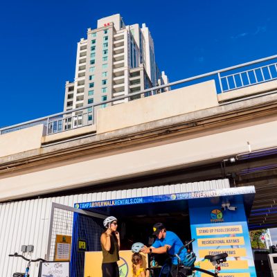 Family renting bikes at a kiosk under a bridge on a sunny day, with tall buildings in the background.
