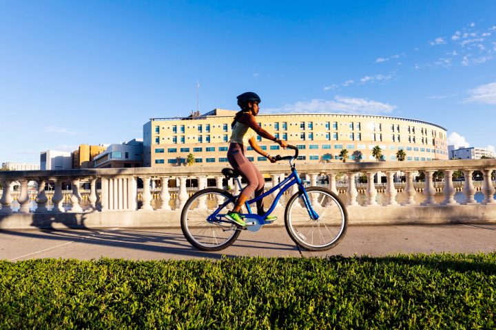 Person rides a blue bike on a sunny day, cityscape and modern building in the background.
