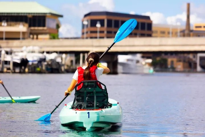 Person kayaking on a calm waterway with buildings and a bridge in the background.