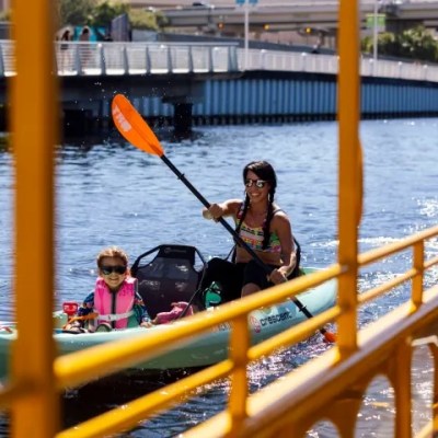 Woman and child kayaking under a yellow bridge on a sunny day.
